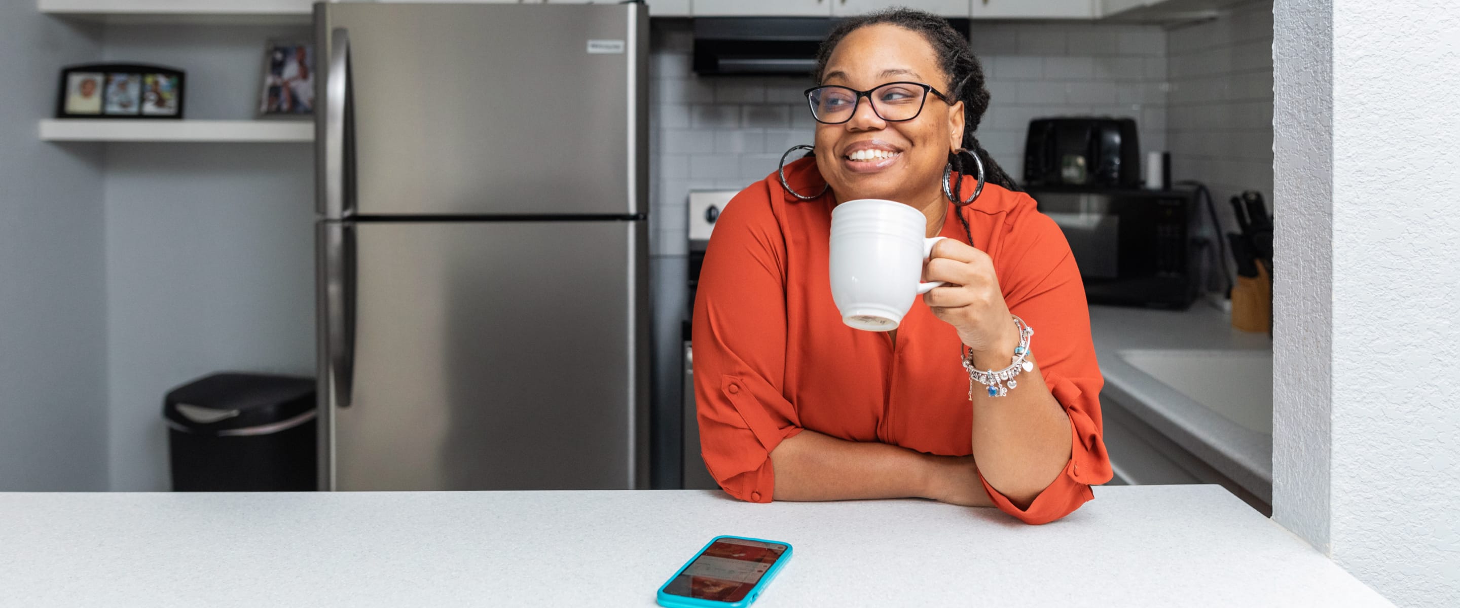 A woman in an orange outfit holding a white mug smiling at a kitchen countertop with a fridge behind her.