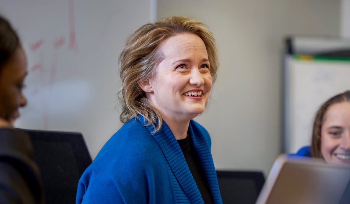 A woman with short hair in a blue suit looking up and smiling in a room with a white board behind her